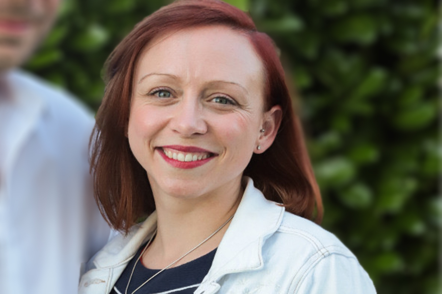 Portraitfoto Jennifer Einolf: rötliche schulterlange Haare, Seitenscheitel, roter Lippenstift, schmale gebogene Augenbrauen, weiße Jeansjacke, rundliches Gesicht, hohe Stirn