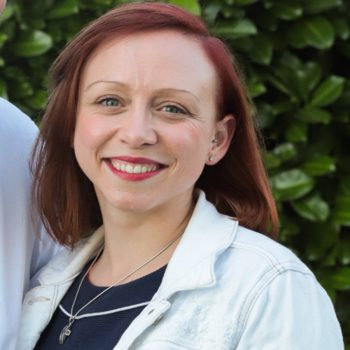 Portraitfoto Jennifer Einolf: rötliche schulterlange Haare, Seitenscheitel, roter Lippenstift, schmale gebogene Augenbrauen, weiße Jeansjacke, rundliches Gesicht, hohe Stirn